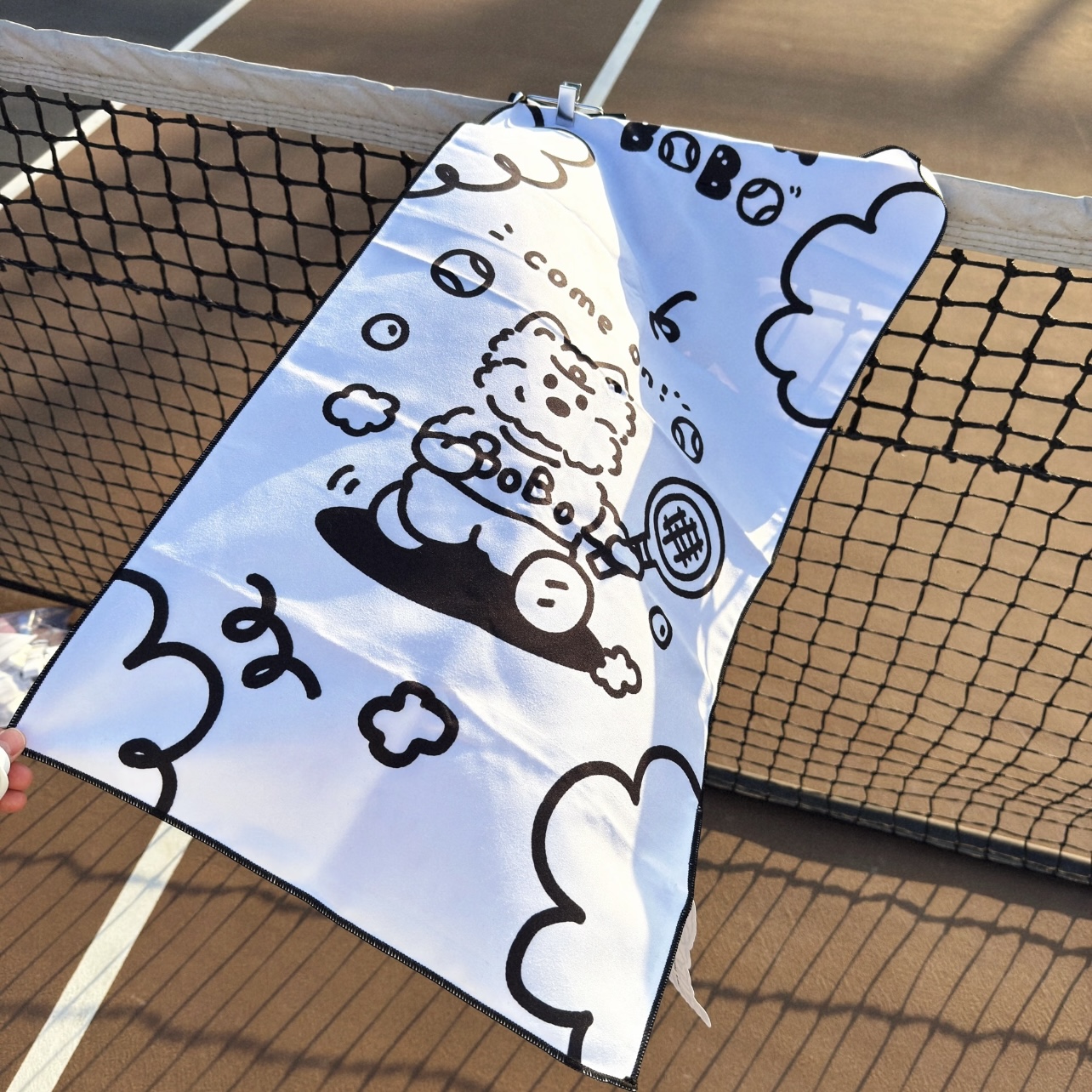 Illustrated black and white custom tennis towel displayed on a net